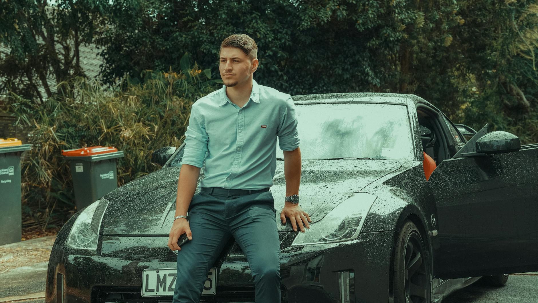 Młody mężczyzna w garniturze oparty o srebrne sportowe Porsche na zewnątrz